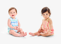 2 young babies sitting on the ground side by side smiling at the camera wearing new rompers.