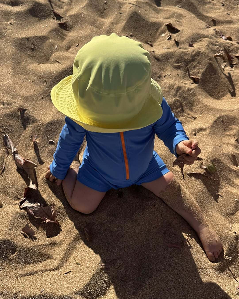 A baby wearing a blue one-piece rash guard and a yellow sun hat from Primary sits on the beach with their head down and legs stretched out.