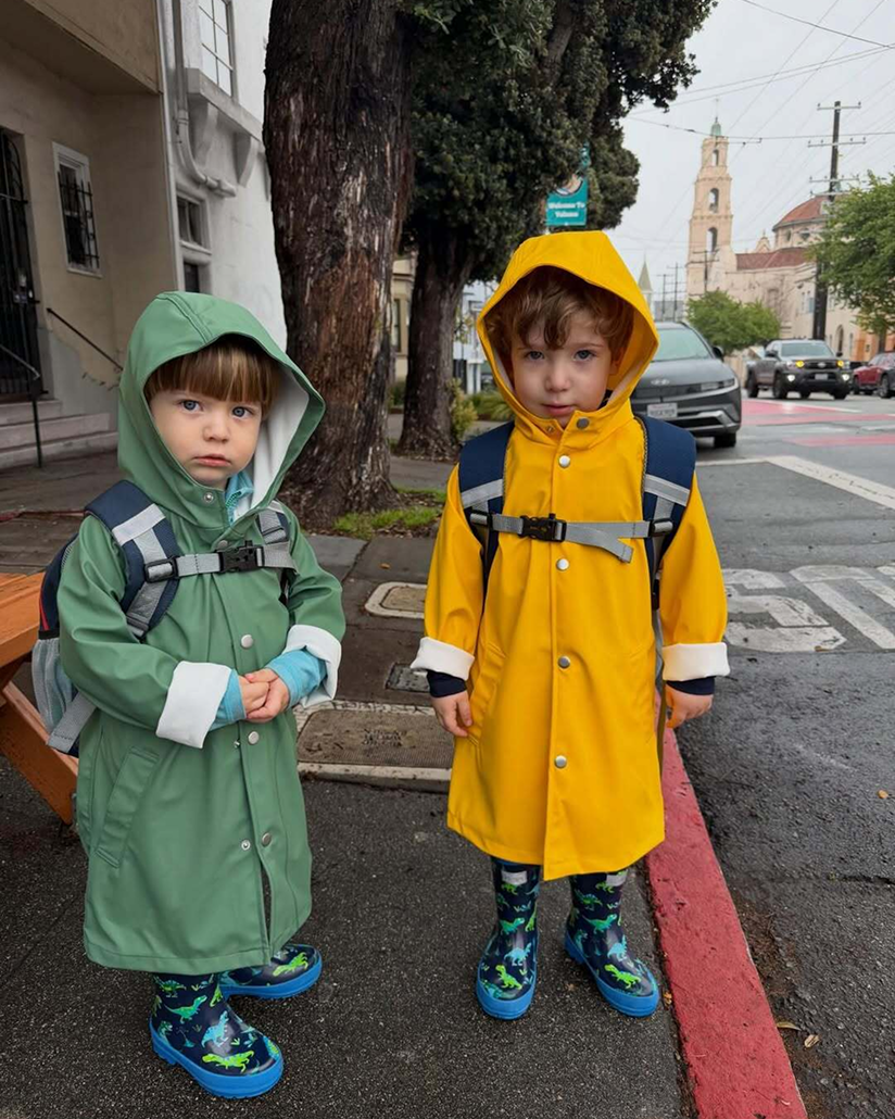 Two kids stand on a wet sidewalk in raincoats from Primary, with rain boots and backpacks—one in green and the other in yellow.