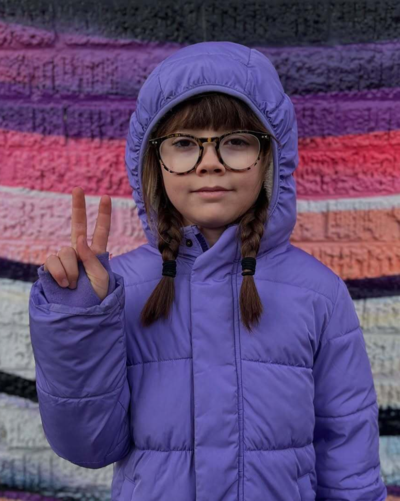 A kid wearing Primary’s purple puffer jacket and glasses flashes a peace sign in front of a colorful striped wall, with a calm expression.