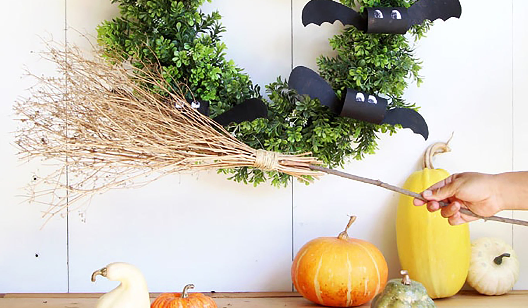 Broomstick made of branches held by hand over table of pumpkins and bat decorations.