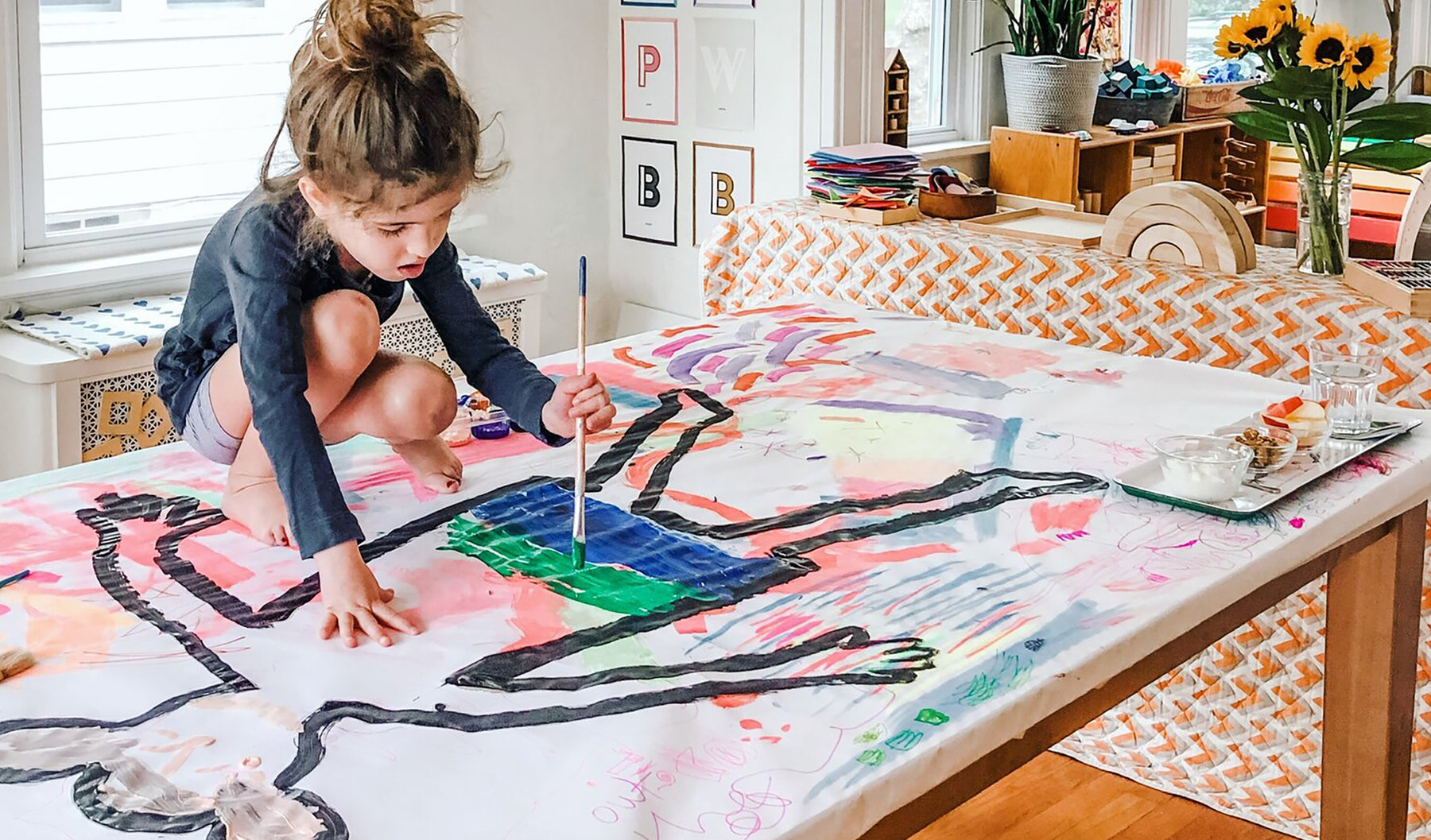 Photo of child on a table, painting a life-size outline of a person.