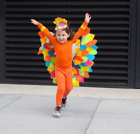 Bird: A child wearing an orange outfit with colorful bird inspired wings and a headdress