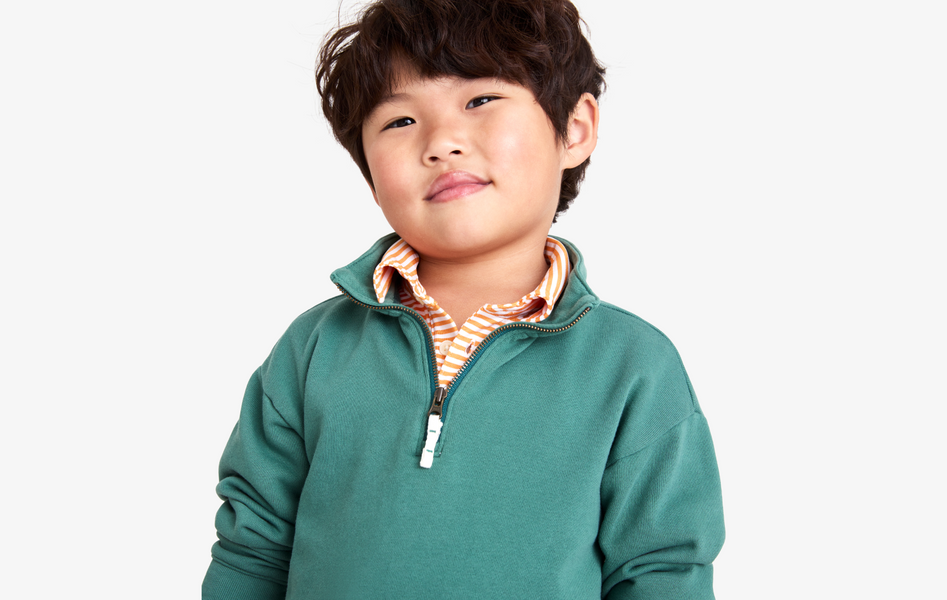 A young child, sitting down and laughing, wearing a green zip-up hooded sweatshirt with a rainbow stripe along the sleeve.