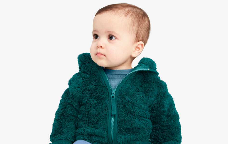 A young baby sitting upright, wearing a spruce color zip up cozy fleece jacket 