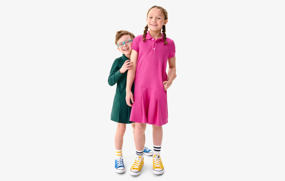 Two young children wearing solid color uniform polo dresses, available with short or long sleeves.