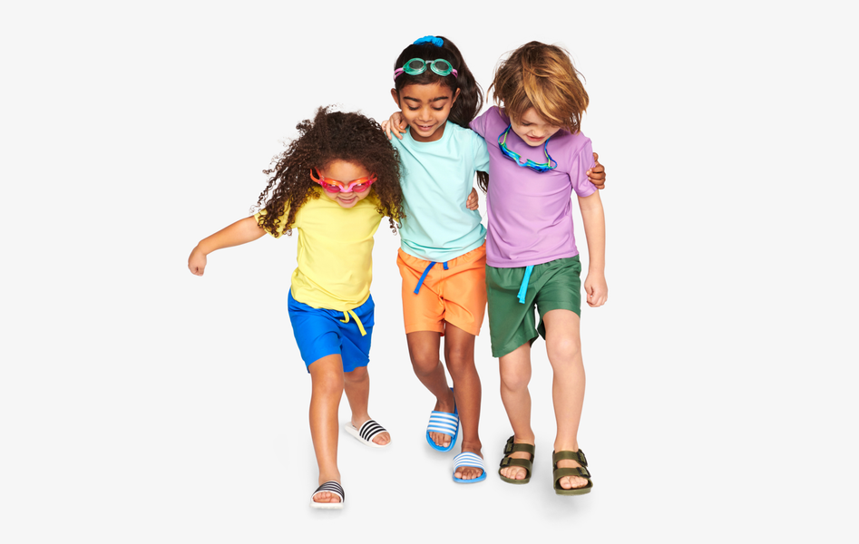 3 children hugging and walking in a line wearing solid long sleeve or short sleeve rash guards with swim trunks and goggles. 