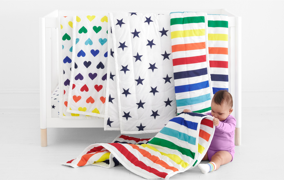 A baby crib draped with different rainbow Primary baby quilts, and a young baby sitting at the foot of crib
