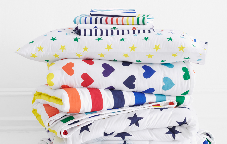A stack of kids sheets, pillows and blankets in rainbow stripes, rainbow stars, and rainbow heart patterns.
