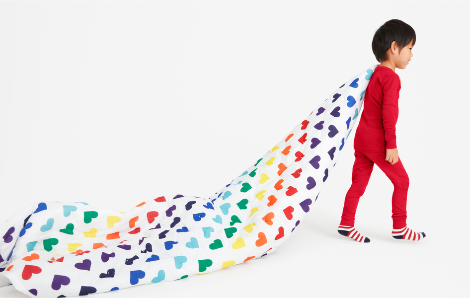 A young child wearing red pajamas, and walking with a rainbow heart print quilt thrown over his shoulder.