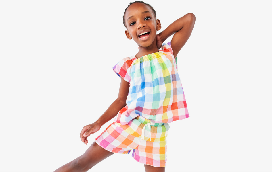 A child smiling at the camera with her hand behind her head wearing our rainbow gingham top and short.