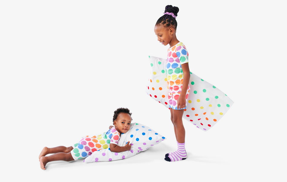 Two children wearing matching short sleeve pajamas with a sunburst rainbow pattern, and pillows with rainbow star pillowcases