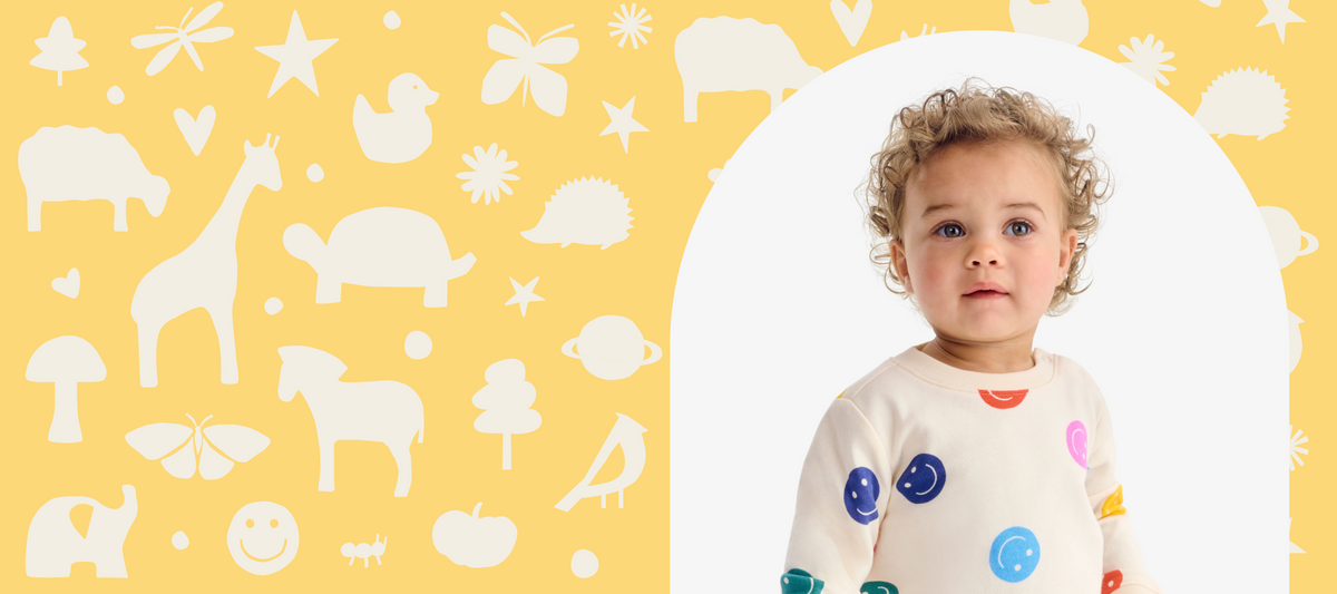 A toddler with curly light brown hair looks directly at the camera, wearing a cream-colored long-sleeve shirt decorated with colorful smiley face and dot patterns. The background is a bright yellow patterned wall with white silhouettes of animals and nature motifs, framed by a white arch on the right.
