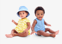 2 babies sitting back to back wearing a yellow dress with bucket hat and a stripe romper.