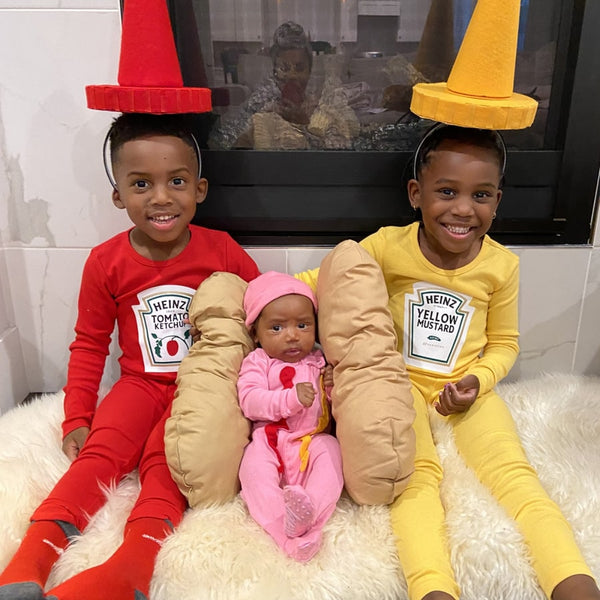 A boy dressed up as a ketchup on the left, a baby wearing pink one-piece in the middle, and a boy dressed up as a mustard mayo sauce on the right.