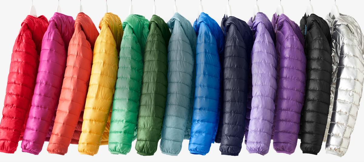 A row of puffer jackets hanging side by side in a rainbow color assortment, including red, pink, orange, yellow, green, blue, navy, purple, black, and silver, against a plain white background.