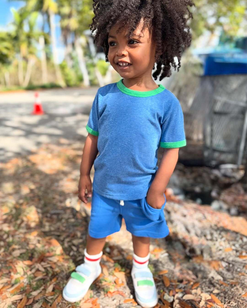 A kid with curly hair stands outdoors, wearing Primary's Kids blue heathered ringer tee and shorts, with left hand in pocket and smiling.