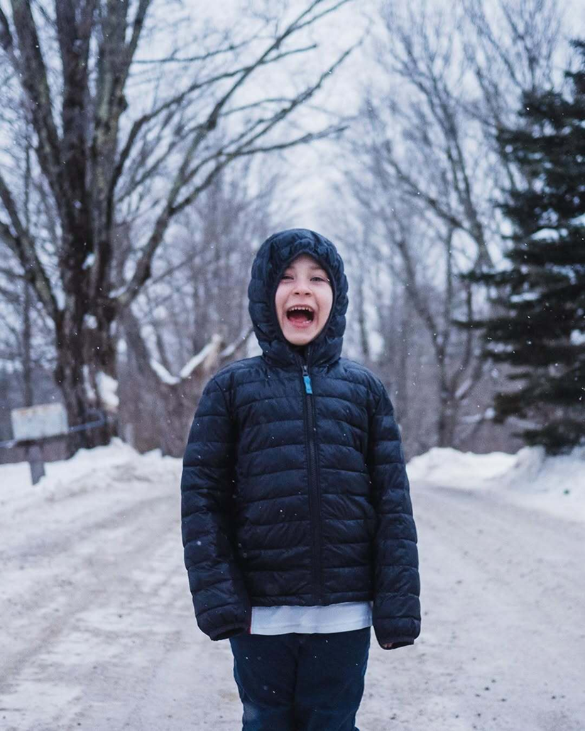 A smiling child wearing Primary’s navy lightweight puffer jacket stands on a snowy path lined with bare trees.