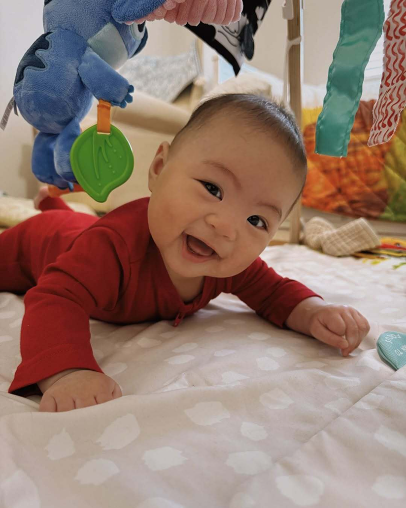 A baby in Primary’s red organic zip romper smiles on a play mat, surrounded by colorful hanging toys.