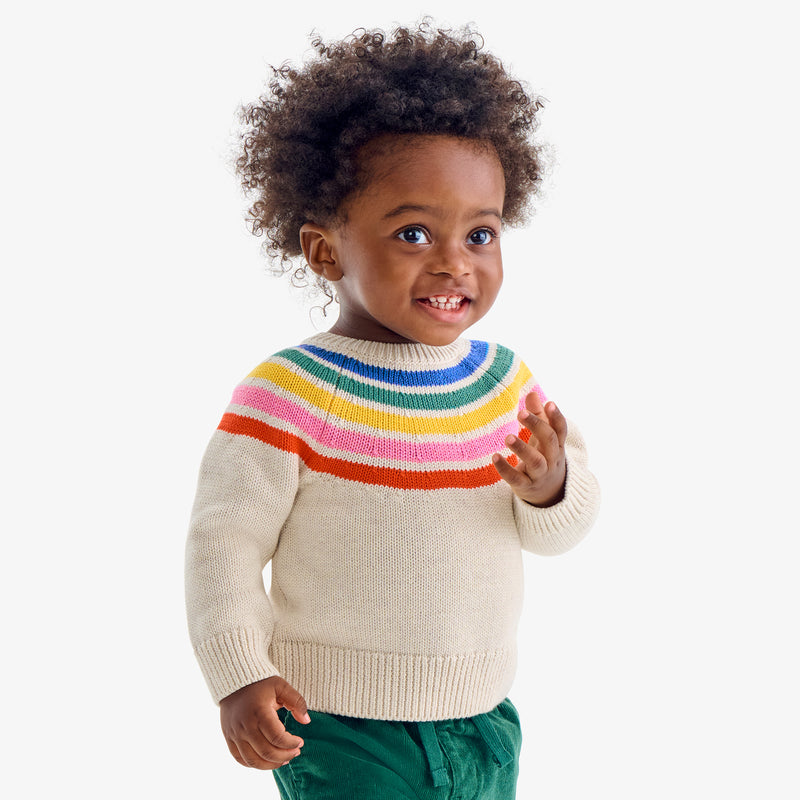 A baby wearing a cream sweater with a rainbow yoke and green corduroy pants