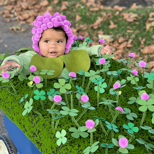 A baby wearing a purple hat and green long sleeve sitting in a cart of handmade clovers.