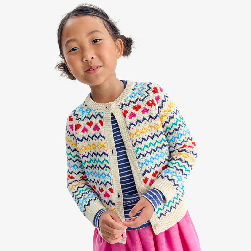 A girl wearing a cream cardigan with a multicolor heart fair isle pattern over a striped top and pink skirt