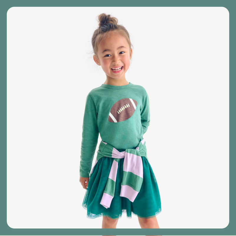 A girl standing and smiling at the camera wearing a green shirt with a football on it with a tut skirt and a shirt tied around her waist 